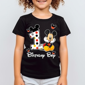 Puede incluir: Camiseta negra con un dise&ntilde;o de Mickey Mouse y el n&uacute;mero 1. El n&uacute;mero 1 tiene un dise&ntilde;o de lunares. El texto "Birthday Boy" est&aacute; escrito en blanco. Mickey Mouse est&aacute; sentado.
