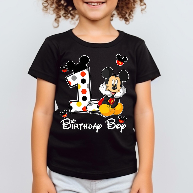 Puede incluir: Camiseta negra con un dise&ntilde;o de Mickey Mouse y el n&uacute;mero 1. El n&uacute;mero 1 tiene un dise&ntilde;o de lunares. El texto "Birthday Boy" est&aacute; escrito en blanco. Mickey Mouse est&aacute; sentado.