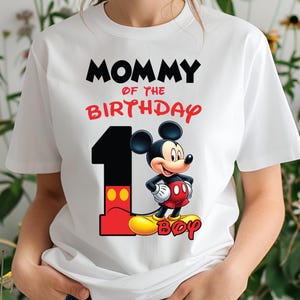 Op de afbeelding: Wit T-shirt met de tekst "MOMMY OF THE BIRTHDAY" in zwart en rood. Een groot nummer "1" staat naast een cartoon Mickey Mouse-personage. Het woord "BOY" staat in het geel onderaan.