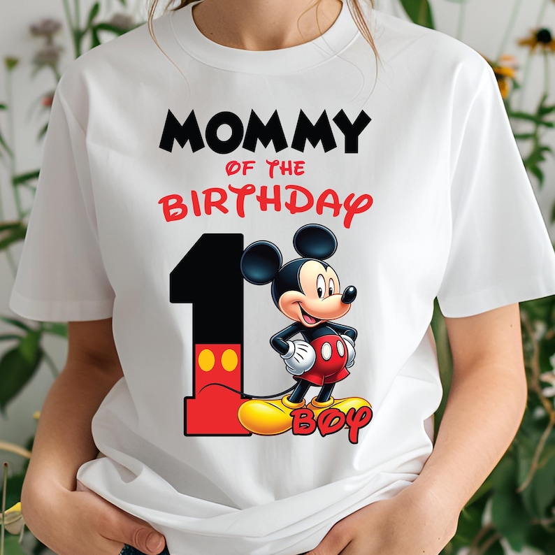 Op de afbeelding: Wit T-shirt met de tekst "MOMMY OF THE BIRTHDAY" in zwart en rood. Een groot nummer "1" staat naast een cartoon Mickey Mouse-personage. Het woord "BOY" staat in het geel onderaan.