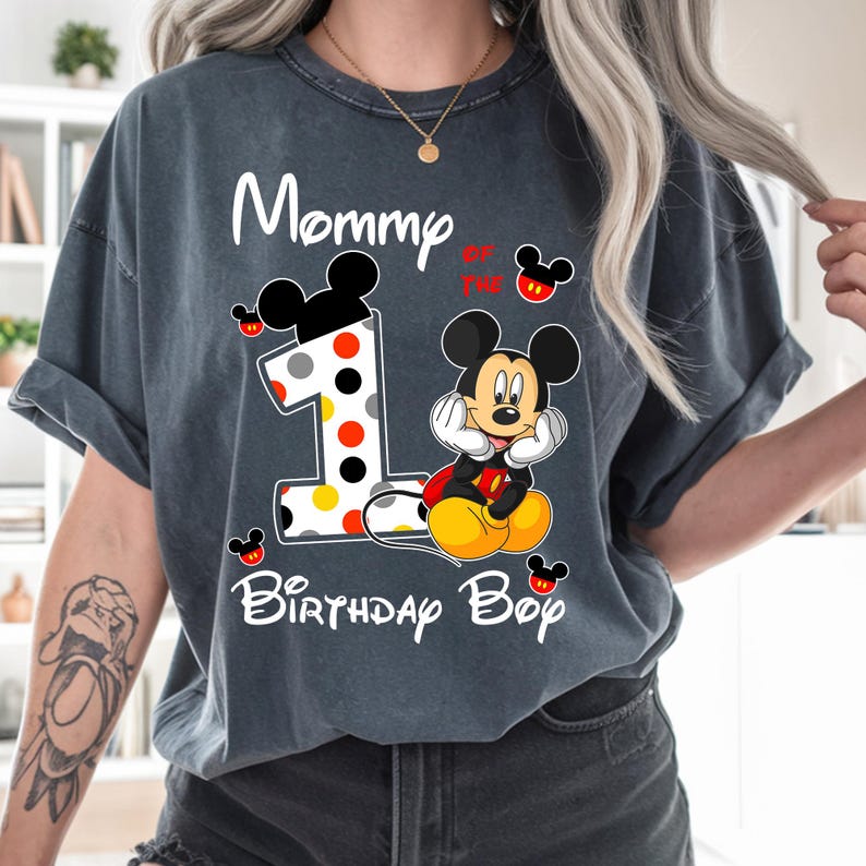 Puede incluir: Camiseta gris oscuro con Mickey Mouse y el texto "Mommy of the 1st Birthday Boy". El n&uacute;mero 1 est&aacute; decorado con lunares. Mickey Mouse est&aacute; en una pose cl&aacute;sica, con las manos bajo la barbilla.