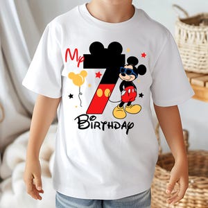 May include: White t-shirt featuring a graphic of Mickey Mouse wearing sunglasses, with the number 7 in red and black, and the words "My Birthday". The design includes stars and a yellow balloon.