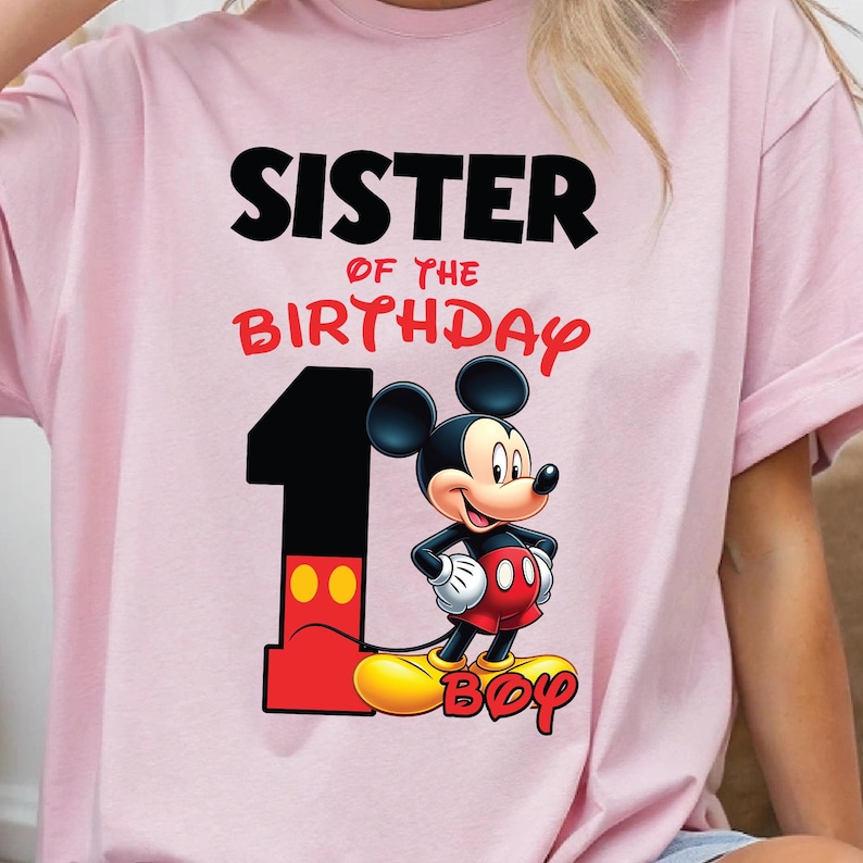 Op de afbeelding: Roze T-shirt met de tekst "SISTER OF THE BIRTHDAY" in zwart en rood. Een afbeelding van Mickey Mouse en het cijfer 1 in zwart en rood staan ook op het shirt. Het woord "BOY" is rood geschreven.