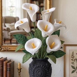 Op de afbeelding: Een gehaakt bloemstuk met witte calla's met gele centra en groene bladeren, gepresenteerd in een donkergrijze vaas. De vaas heeft een gestructureerd, patroonontwerp. De opstelling staat op een houten oppervlak.