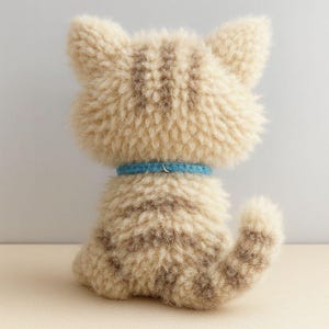 Puede incluir: Un juguete de peluche con forma de gato visto desde atr&aacute;s, con un patr&oacute;n de rayas beige claro y marr&oacute;n. El gato tiene un collar azul con un cierre plateado. El pelaje del juguete es texturizado y esponjoso, con una peque&ntilde;a cola curvada hacia un lado.
