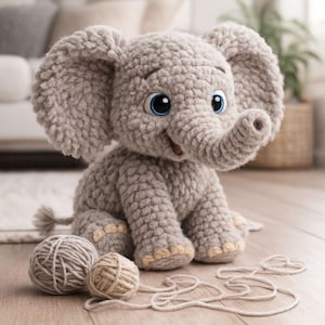 以下が含まれることがあります： 大きな耳、鼻、大きな青い目を持つ、手作りの灰色の編み物象。 象は、2つの毛糸玉の隣の木製の表面に座っています。 象の足と耳は薄いタン色です。