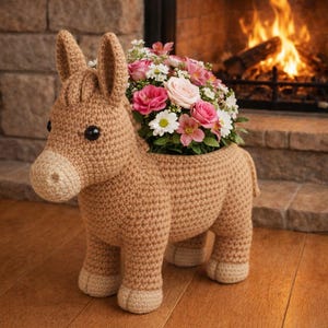 Op de afbeelding: Een handgemaakte, gehaakte ezel-bloempot in beige met een bloemstuk. De ezel heeft zwarte ogen en een lichtbeige snuit en poten. De bloempot is gevuld met roze, witte en rode bloemen. Op de achtergrond is een open haard te zien.