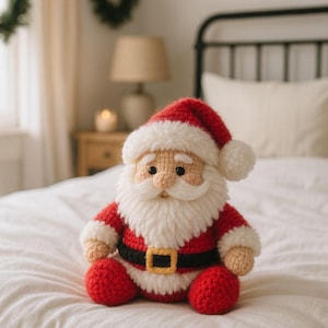 Puede incluir: Muñeco de Papá Noel tejido a ganchillo, con gorro y traje rojos, ribete blanco, barba blanca esponjosa y cinturón negro con hebilla dorada. El muñeco está sobre una colcha blanca. Decoración navideña.