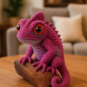 Peut inclure: Un caméléon au crochet fait à la main, dans des tons de rose et de violet, perché sur une base en bois. Le reptile présente de grands yeux expressifs avec des détails orange et noirs. Ce jouet en peluche est un objet décoratif unique.