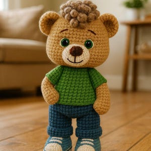 Puede incluir: Un oso de peluche hecho a mano con ganchillo, que lleva una camiseta verde, vaqueros azules y zapatillas azules. El oso tiene una nariz marrón, ojos verdes y un mechón de pelo castaño. Está de pie sobre un suelo de madera.