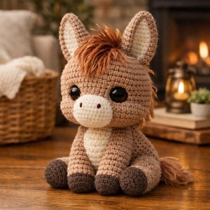 May include: A handmade crochet donkey toy in shades of brown, tan, and cream. The donkey has large black eyes, a white muzzle, and a tuft of brown yarn for a mane. The toy is sitting on a wooden surface.