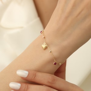 Può includere: Un delicato braccialetto d'oro con un ciondolo a forma di cuore, piccole perline e accenti color rubino. Il braccialetto è disegnato con una catena sottile e viene mostrato su un polso.