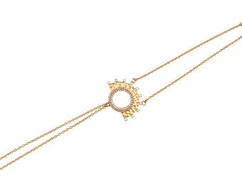 Puede incluir: Una pulsera de cadena dorada con un colgante central semicircular de textura martillada. El colgante tiene un recorte circular rodeado de pequeñas piedras claras, con piedras claras adicionales a lo largo del borde curvo exterior.