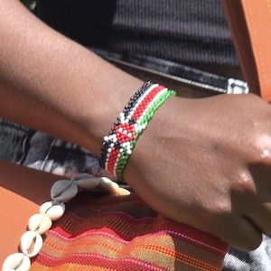 May include: A colourful beaded bracelet, featuring the Kenyan flag's colours: black, red, green, and white. The bracelet is worn on a wrist. A striped textile and a seashell necklace are also visible.