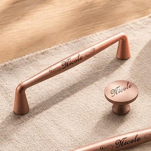 May include: A collection of rose gold cabinet hardware including three elongated bar pulls and three round knobs. Each piece features a smooth metallic finish and the name Nicole engraved in a cursive script on the front surface of the hardware.