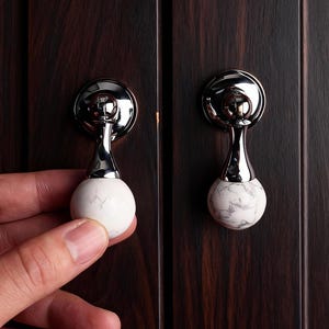Könnte beinhalten: Schrankknöpfe mit polierter Chromoberfläche und einer weißen Kugel in Marmoroptik. Die Knöpfe sind an einer dunkelbraunen Holzschranktür montiert. Einer der Knöpfe wird von einer Hand gehalten.