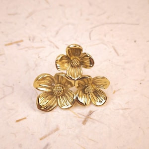 Puede incluir: Un broche de metal en tono dorado que presenta tres flores tridimensionales dispuestas en racimo. Cada flor tiene cuatro pétalos redondeados con superficies texturizadas y estriadas, y un pequeño centro granular elevado. El metal tiene un acabado pulido.