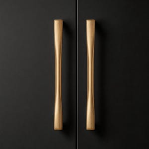 Può includere: Primo piano di due maniglie dorate per armadi contro un'anta nera. Le maniglie hanno un design elegante e allungato con una forma leggermente a clessidra. L'hardware aggiunge un tocco di eleganza all'armadio scuro.