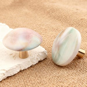 May include: Two decorative mushroom-shaped cabinet knobs. One knob is mushroom-shaped with a pink, white, and blue top and a gold-colored stem. The other knob is oval-shaped with a white, green, and pink top and a gold-colored stem.