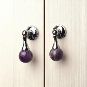 May include: Two cabinet pulls with a polished chrome finish and a spherical, deep purple stone accent. The pulls are mounted on a white cabinet door. The pulls have a decorative design.