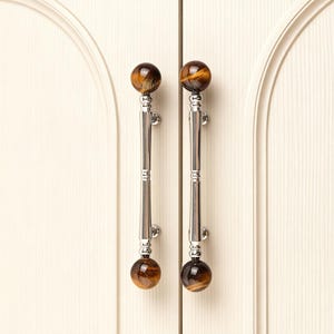 May include: Two cabinet pulls with a polished chrome finish and tiger's eye stone spheres. The pulls are vertically oriented, with a decorative design in the center. The cabinet doors are a light cream color.