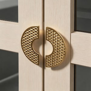 May include: Close-up of two gold-colored cabinet door handles. Each handle is a half-circle shape with a textured, scale-like pattern. The handles are mounted on light-colored wooden cabinet doors.