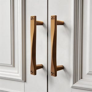 Può includere: Due maniglie per mobili color bronzo con un design rettangolare nella parte superiore e inferiore. Le maniglie hanno una forma curva e attorcigliata e sono montate su un'anta bianca. Le maniglie sono lunghe circa 15 cm.
