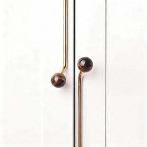 Puede incluir: Primer plano de dos tiradores de puerta de armario. Cada tirador presenta un vástago curvo de color dorado con un pomo redondo de piedra ojo de tigre marrón y dorado. Los tiradores están montados en una puerta de armario blanca.