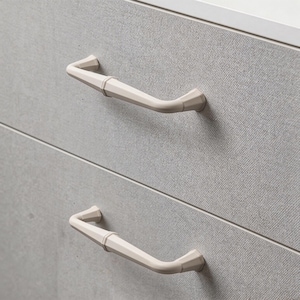Puede incluir: Primer plano de dos tiradores de armario blanquecinos con un diseño inspirado en el bambú. Los tiradores están montados en una puerta de armario texturizada de color gris claro. Los tiradores tienen forma curva.