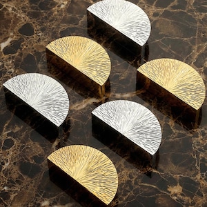 May include: Six half-moon cabinet pulls in gold and silver. Each pull has a textured, sunburst design. The hardware is arranged on a brown and tan marble surface, creating a contrast of colours and textures.