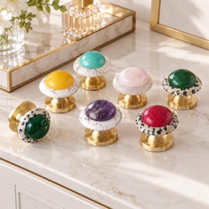 May include: A collection of seven decorative cabinet knobs featuring polished brass bases and circular tops. Each knob has a unique gemstone-like center in colors including turquoise, yellow, pink, dark green, purple, and red, set in stone.