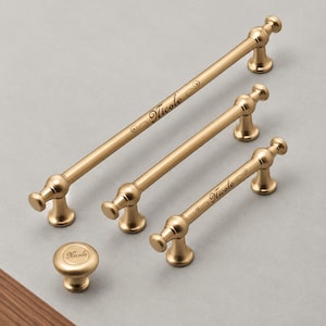 May include: Three gold-toned metal cabinet pulls of varying lengths and one matching round cabinet knob. Each piece features a matte finish and the name Nicole engraved in a cursive script with decorative flourishes on the handle surfaces.