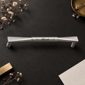 May include: A brushed silver metal cabinet handle with a tapered design and two cylindrical mounting posts. The center of the handle features the engraved name Nicole in a cursive script font, flanked by decorative scrollwork on both sides.