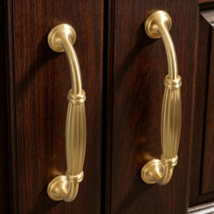 May include: Two gold-colored cabinet handles with a curved design. The handles are mounted on a dark brown wooden cabinet door. The handles have a decorative knot detail and a fluted center section.