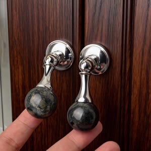 Könnte beinhalten: Zwei Schrankgriffe mit polierter Nickeloberfläche und einer dunkelgrünen und schwarzen Marmorkugel. Die Griffe sind an einer dunklen Holzschranktür befestigt. Die Griffe sind rund mit einem dekorativen Design.