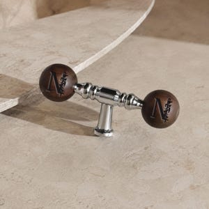 May include: A T-shaped cabinet handle featuring a polished chrome centre post and two dark brown wooden spheres on either end. Each wooden sphere is engraved with the letter N accompanied by a decorative floral flourish design on the side.