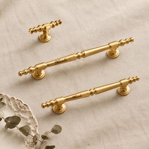 May include: Two long gold-toned metal cabinet pulls and one small matching gold-toned knob. Each pull features a central cylindrical bar with decorative beaded ends and the name Nicole engraved in a script font on the centre surface.