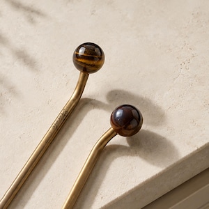 May include: Two metal stirring sticks with a matte gold finish, each topped with a polished spherical gemstone. One stick features the name Nicole engraved in script on the shaft. The stones exhibit brown and gold banded patterns.