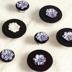 Pode incluir: Puxadores decorativos para armários com base circular castanho escuro e design floral 3D. As flores são em tons de branco e lavanda. Os puxadores estão dispostos sobre um fundo branco texturizado.