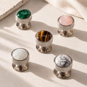 May include: Five circular cabinet knobs with silver-toned, textured metal bases. Each knob features a unique stone inlay: green malachite, pale pink quartz, brown tiger's eye, white moonstone, and white marble with grey veining. Each has the name Nicole.