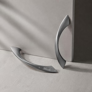 May include: Two matte grey metal cabinet handles with a curved, ergonomic design. One handle is positioned vertically, while the other lies horizontally and features the name Nicole engraved in a cursive script with decorative flourishes on its surface.