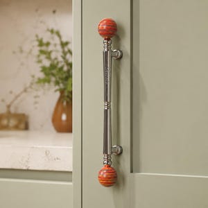 May include: A vertical cabinet handle featuring a polished silver-tone metal bar with engraved text and two spherical ends. Each sphere displays a vibrant, multi-colored horizontal striped pattern in shades of orange, red, and yellow.