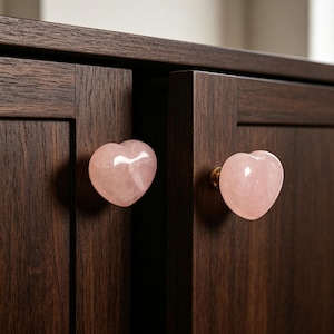 Peut inclure: Deux boutons de placard en forme de cœur en quartz rose fixés à une armoire en bois brun foncé. Les boutons sont de couleur rose tendre et ont une surface lisse et polie. Les portes de l'armoire sont fermées et ont un design simple et moderne.