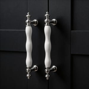 Puede incluir: Dos tiradores de armario de cerámica blanca con base y detalles plateados. Los tiradores tienen una forma curva y alargada y están montados en una puerta de armario oscura, posiblemente negra.