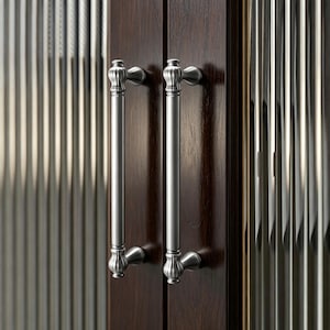 Könnte beinhalten: Zwei Türgriffe für Schränke aus gebürstetem Nickel mit dekorativem Design. Die Griffe sind an einer dunkelbraunen Holzschranktür mit einer geriffelten Glasscheibe angebracht.