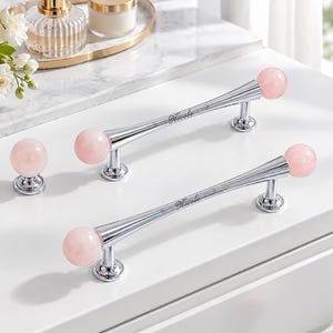 May include: Two silver-toned metal cabinet handles and one matching round knob, each featuring polished pale pink spherical stone accents at the ends. The handles have a tapered design with the script text Nicole engraved on the central bar.