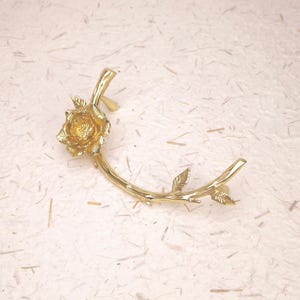 Pode incluir: Um ear cuff dourado em forma de ramo curvo com uma rosa detalhada numa extremidade. A superfície metálica é polida e refletora, apresentando pétalas texturizadas na flor e pequenas folhas ao longo do caule do ramo.