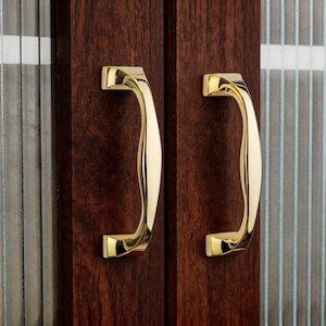 以下が含まれることがあります： 濃い茶色の木製ドアに取り付けられた、磨かれた金色のドアハンドル2つのクローズアップ。ハンドルは湾曲した人間工学に基づいたデザインで、光沢のある仕上がりです。ドアには、垂直線のあるガラスパネルが付いています。