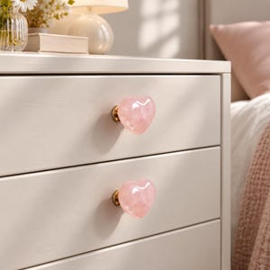 May include: Two heart-shaped drawer knobs made of translucent pale pink stone, each attached to a circular gold-toned metal base. The knobs are mounted on the front of a light cream-colored wooden dresser with two horizontal drawers.