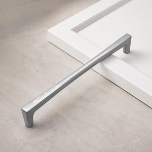 May include: A matte silver cabinet pull handle with a rectangular profile and tapered ends. The surface features the engraved name Nicole in a cursive script font, accompanied by delicate swirling decorative flourishes on either side of the text.